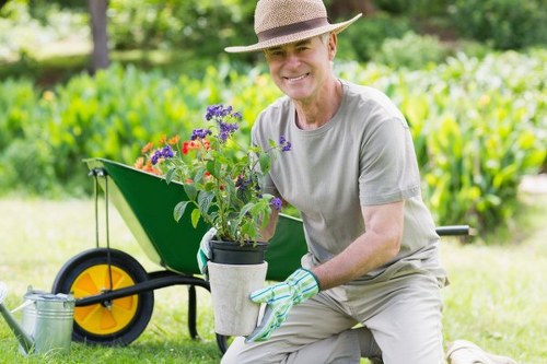 A smiling elderly man wearing a straw hat, light grey t-shirt, and gardening gloves is kneeling on well-maintained grass in a backyard garden. He is holding a small potted flowering plant with purple and red blooms, positioned towards the camera. Behind him, there is a bright green wheelbarrow with yellow and black wheels, containing garden tools and a watering can, indicating active garden maintenance. The background features lush, dense foliage and trees, suggesting a spacious outdoor setting typical of a suburban garden in New Cross. The area showcases a neatly edged lawn, with a paved pathway or patio section partially visible, framed by a mix of shrubs and small trees. The scene is illuminated by natural daylight, creating a vibrant and inviting outdoor environment suitable for gardening services focused on lawn care, shrub trimming, and general garden upkeep in the local area of New Cross, London.