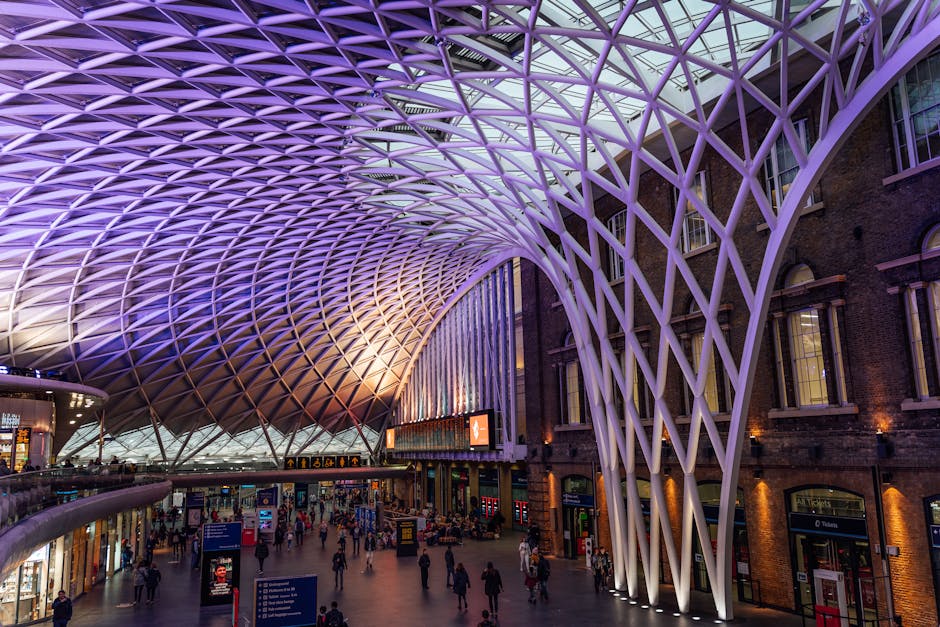 A spacious train station interior featuring a striking, modern architectural canopy with an intricate lattice design illuminated in purple and white lighting. The high, curved ceiling structure is sup